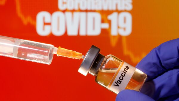 A small bottle labeled with a Vaccine sticker is held near a medical syringe in front of displayed Coronavirus COVID-19 words in this illustration taken April 10, 2020. - Sputnik International