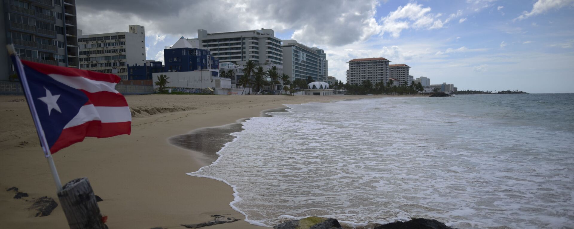 A Puerto Rican flag flies on an empty beach at Ocean Park, in San Juan, Puerto Rico, Thursday, May 21, 2020. Puerto Rico is cautiously reopening beaches, restaurants, churches, malls, and hair salons under strict conditions as the U.S. territory emerges from a two-month lockdown despite dozens of new coronavirus cases reported daily.  - Sputnik International, 1920, 03.02.2021