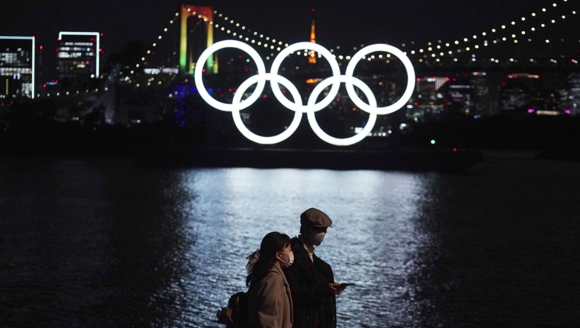 A man and woman walk past the Olympic rings floating in the water in the Odaiba (artificial island) section on Tuesday, 1 December 2020, in Tokyo. The Olympic Symbol was reinstated after it was taken down for maintenance ahead of the postponed Tokyo 2020 Olympics. - Sputnik International, 1920, 20.07.2021