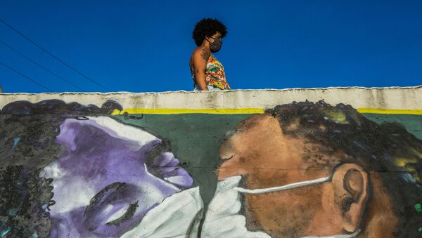  In this file photo taken on April 15, 2020 a woman wearing a face mask stands above a graffiti by artist Marcos Costa, or Spray Cabuloso, at the entrance of the Solar de Unhao favela in Salvador, Bahia state, Brazil. - Sputnik International
