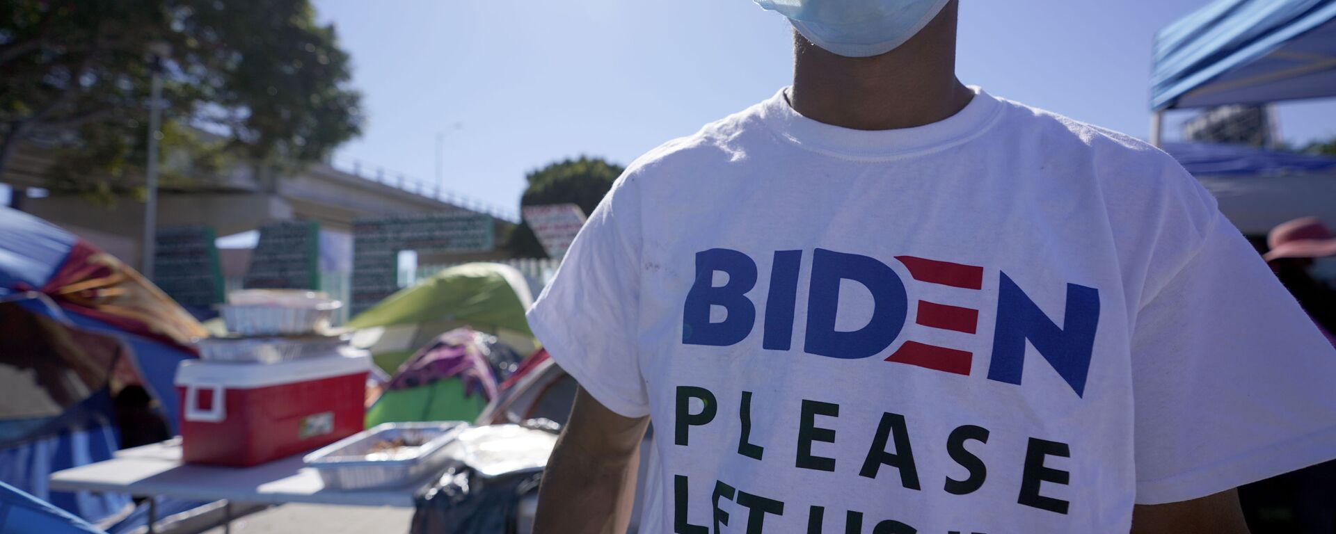 A Honduran man seeking asylum in the United States wears a shirt that reads, Biden please let us in, as he stands among tents that line an entrance to the border crossing, Monday, March 1, 2021, in Tijuana, Mexico. - Sputnik International, 1920, 28.12.2022