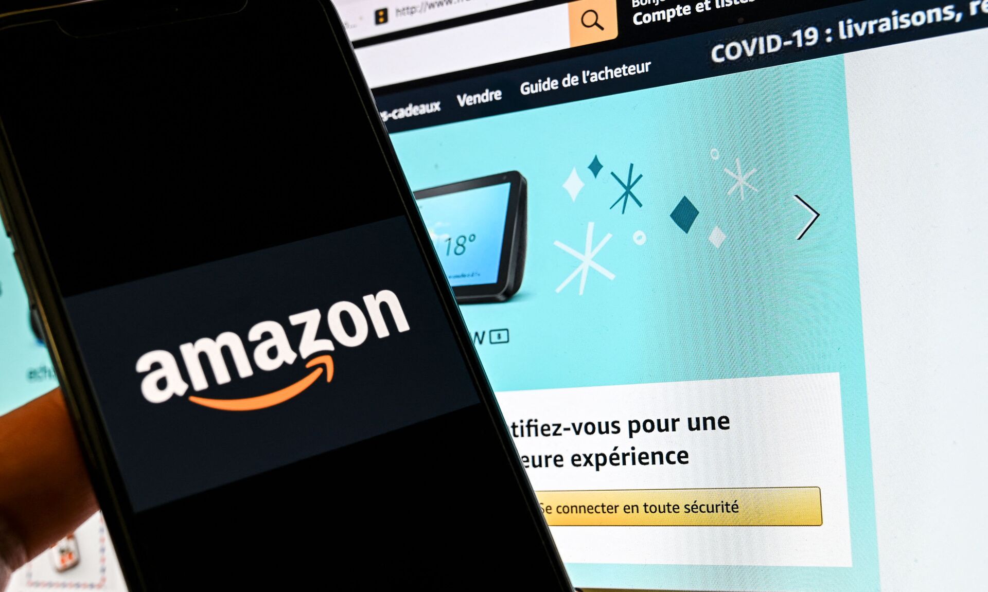 In this photograph taken on November 18, 2020 in Lille, a person poses with a smartphone showing an Amazon logo, in front of a computer screen displaying the home page of Amazon France sales website. - Sputnik International, 1920, 03.12.2021