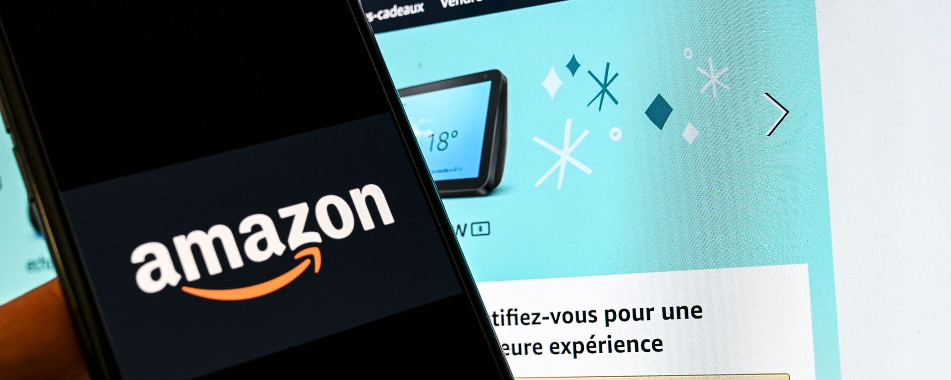 In this photograph taken on November 18, 2020 in Lille, a person poses with a smartphone showing an Amazon logo, in front of a computer screen displaying the home page of Amazon France sales website. - Sputnik International, 1920, 30.07.2021