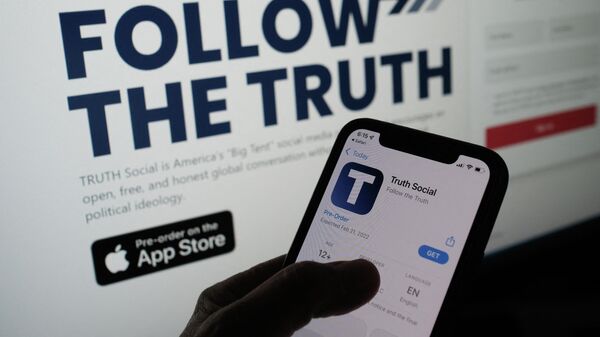 This illustration photo shows a person checking the app store on a smartphone for Truth Social, with it's website on a computer screen in the background, in Los Angeles, October 20, 2021. - Former US president Donald Trump announced plans on October 20 to launch his own social networking platform called TRUTH Social, which is expected to begin its beta launch for invited guests next month. The long-awaited platform will be owned by Trump Media & Technology Group (TMTG), which also intends to launch a subscription video on-demand service that will feature non-woke entertainment programming, the group said in a statement. - Sputnik International