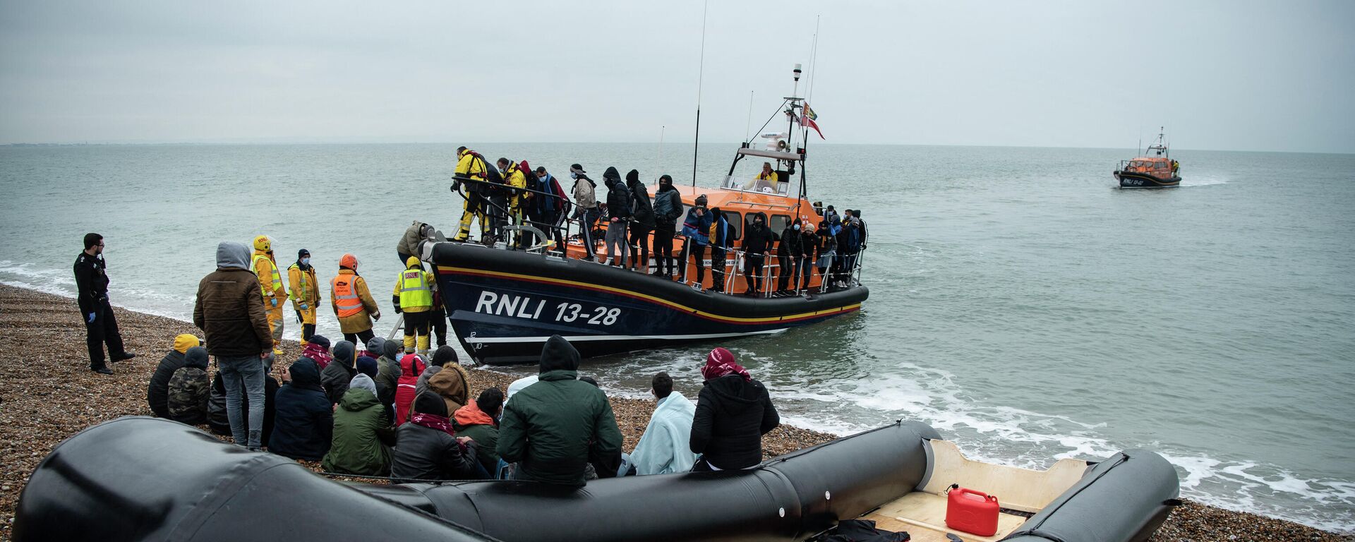 Migrants sit beside a boat used to cross the English Channel as more migrants are helped ashore from a RNLI (Royal National Lifeboat Institution) lifeboat at a beach in Dungeness, on the south-east coast of England, on November 24, 2021 - Sputnik International, 1920, 05.03.2022