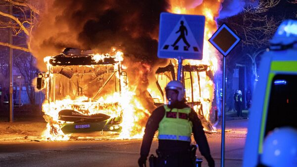 An anti-riot police officer stands next to a city bus burning in Malmo late April 16, 2022. - The unrest in Malmo has continued after Rasmus Paludan, party leader of the Danish right-wing extremist party Tight Course, held a demonstration on April 16, 2022 at Skanegarden near the Oresund Bridge. (Photo by Johan NILSSON / TT NEWS AGENCY / AFP)  - Sputnik International