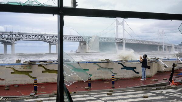 A man standing amongst debris on a road is seen beyond the broken windows (foreground) of a convenience store, while taking photos of waves in front of the Gwangandaegyo or Diamond Bridge (back), after Typhoon Hinnamnor passed through Busan on September 6, 2022 - Sputnik International