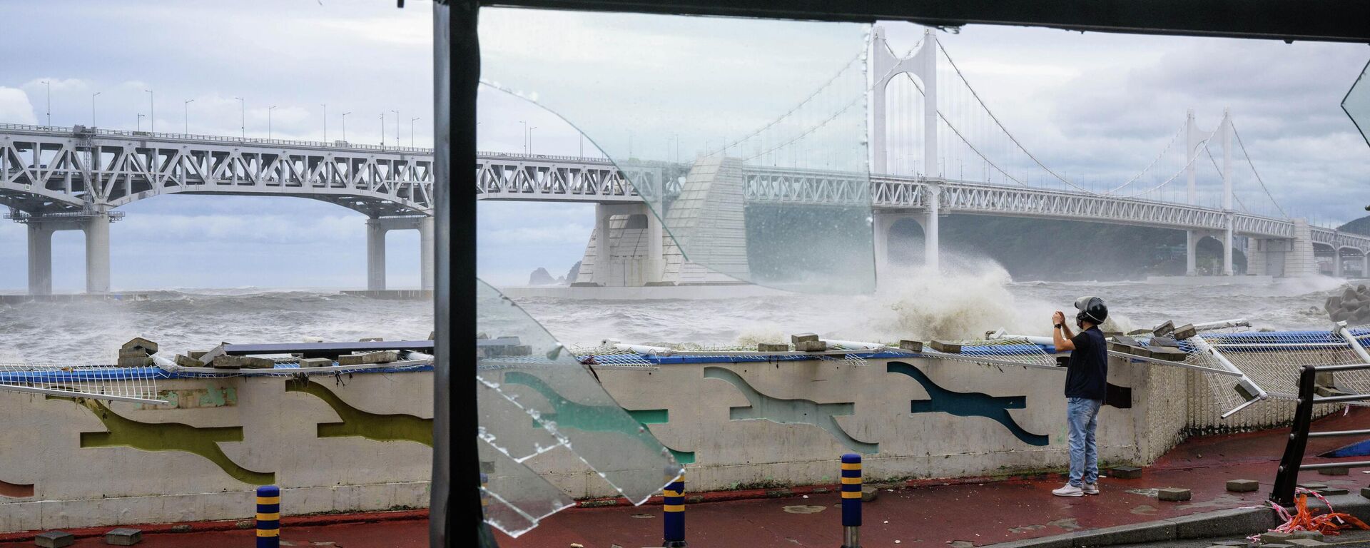 A man standing amongst debris on a road is seen beyond the broken windows (foreground) of a convenience store, while taking photos of waves in front of the Gwangandaegyo or Diamond Bridge (back), after Typhoon Hinnamnor passed through Busan on September 6, 2022 - Sputnik International, 1920, 06.09.2022