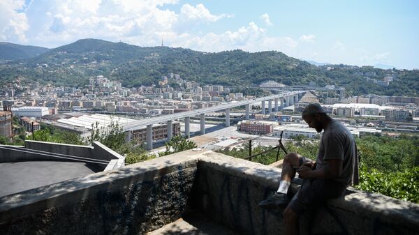 A man checks his phone as he sits overlooking Genoa with the Geneo-San Giorgio Bridge (Ponte Genova-San Giorgio) in the background on 7 July, 2022 - Sputnik International