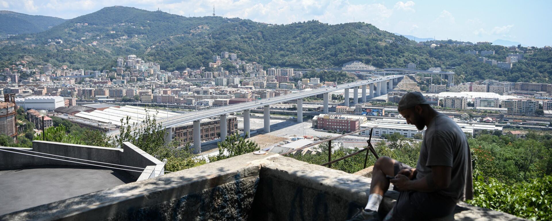 A man checks his phone as he sits overlooking Genoa with the Geneo-San Giorgio Bridge (Ponte Genova-San Giorgio) in the background on 7 July, 2022 - Sputnik International, 1920, 02.12.2022