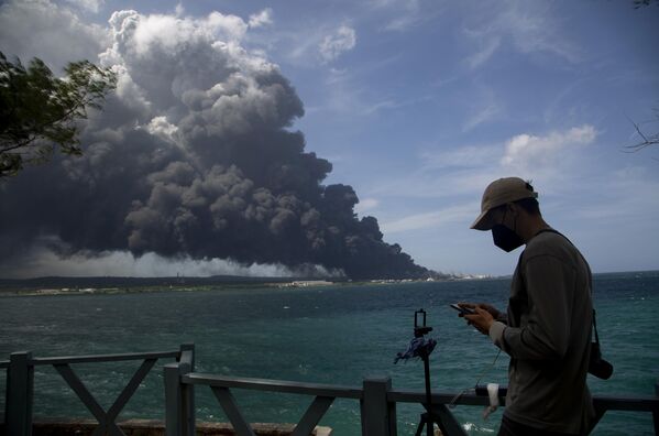 A man types on his cellphone near a huge plume of smoke rising from the Matanzas Supertanker Base as firefighters work to quell the blaze which began during a thunderstorm in Matanzas, Cuba, Monday, Aug. 8, 2022. Cuban authorities say lightning struck a crude oil storage tank at the base, sparking a fire that ignited four explosions. - Sputnik International