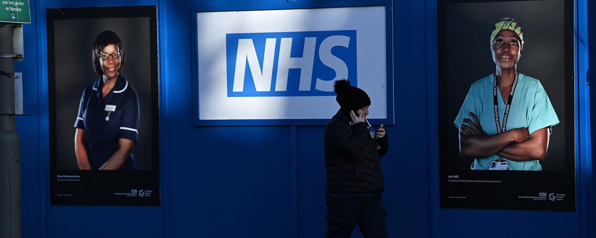 Pedestrians walk past images of workers of Britain's National Health Service (NHS) fixed to hoardings outside a temporary field hospital, set up in the grounds of St George's Hospital in Tooting, south London on January 5, 2022 - Sputnik International, 1920, 02.01.2023