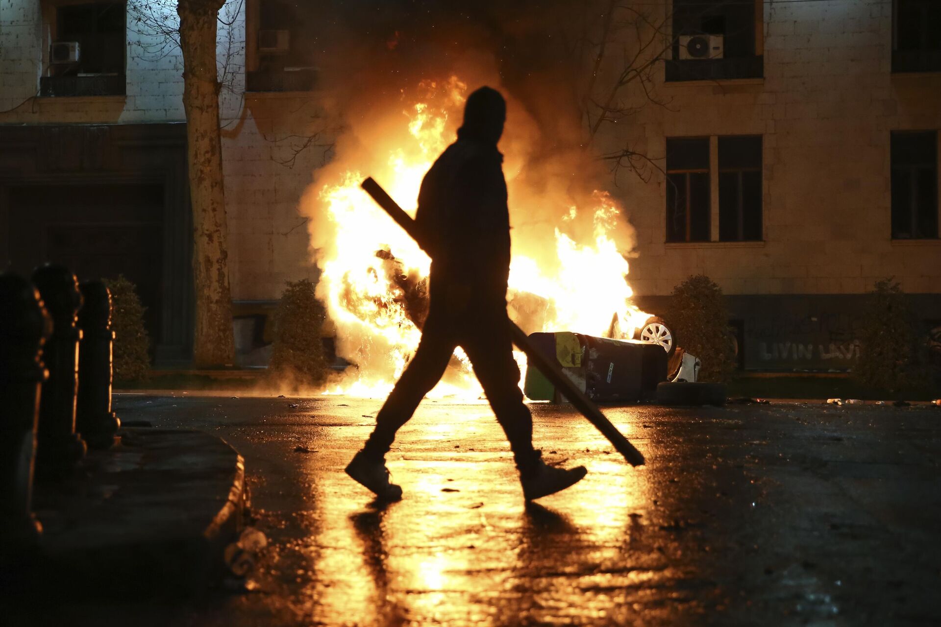 A man with a stick in hand walks past a burning police car not far from the Georgian parliament building in Tbilisi, Georgia, Thursday, March 9, 2023. - Sputnik International, 1920, 16.05.2024