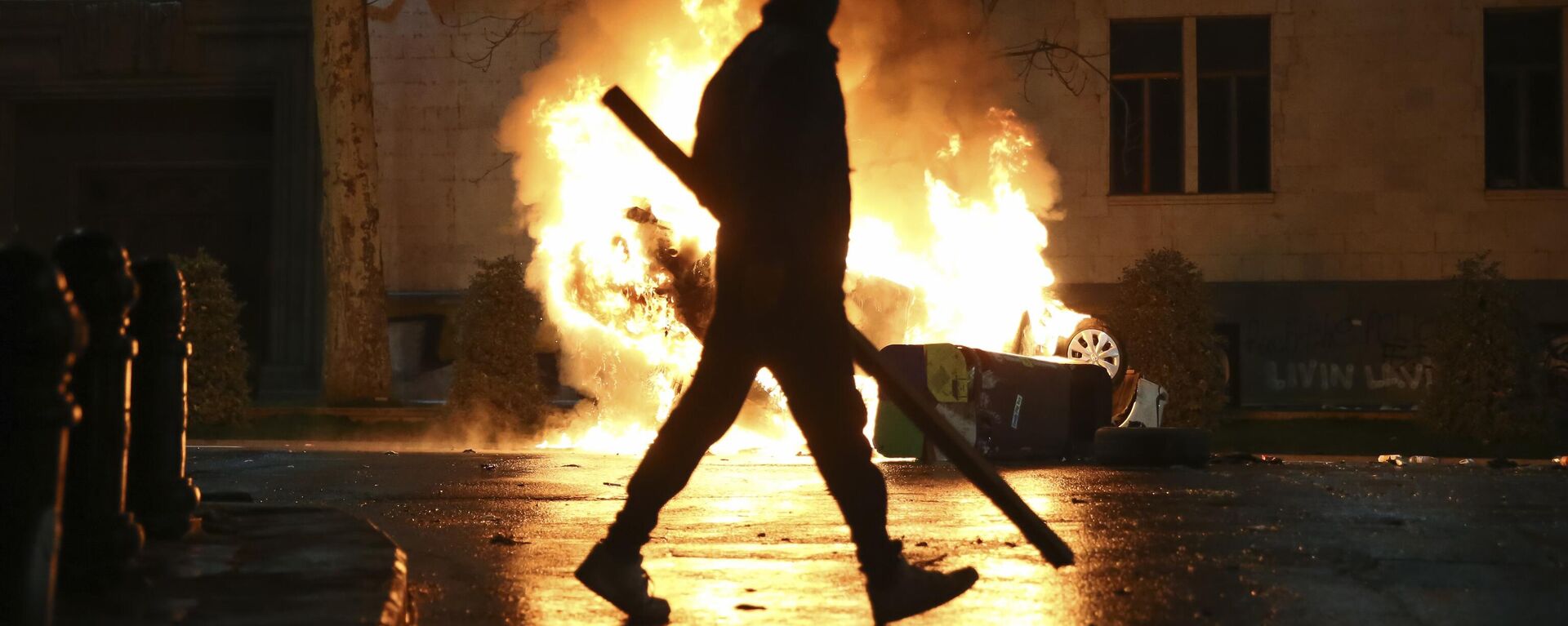 A man with a stick in hand walks past a burning police car not far from the Georgian parliament building in Tbilisi, Georgia, Thursday, March 9, 2023. - Sputnik International, 1920, 21.05.2024