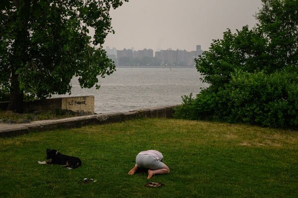 A person stretches before the New York City skyline and East River, both shrouded in smoke, in Brooklyn on June 6, 2023. Smoke from hundreds of wildfires blazing in eastern Canada has drifted south. According to the Canadian Interagency Forest Fire Center, hundreds of wildfires were burning in Canada on June 6, 2023, as fires have been breaking out across the country in recent weeks. The agency reported that over 150 active blazes were present across the province of Quebec alone. - Sputnik International
