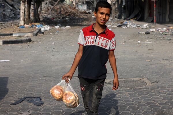 A young boy carries bags of bread as he walks in front of a building damaged by strikes in Gaza City. - Sputnik International