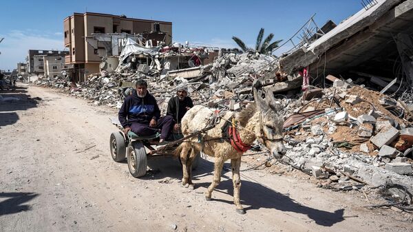 Two men sit in a donkey-drawn cart moving past the rubble of a destroyed building in Khan Yunis - Sputnik International