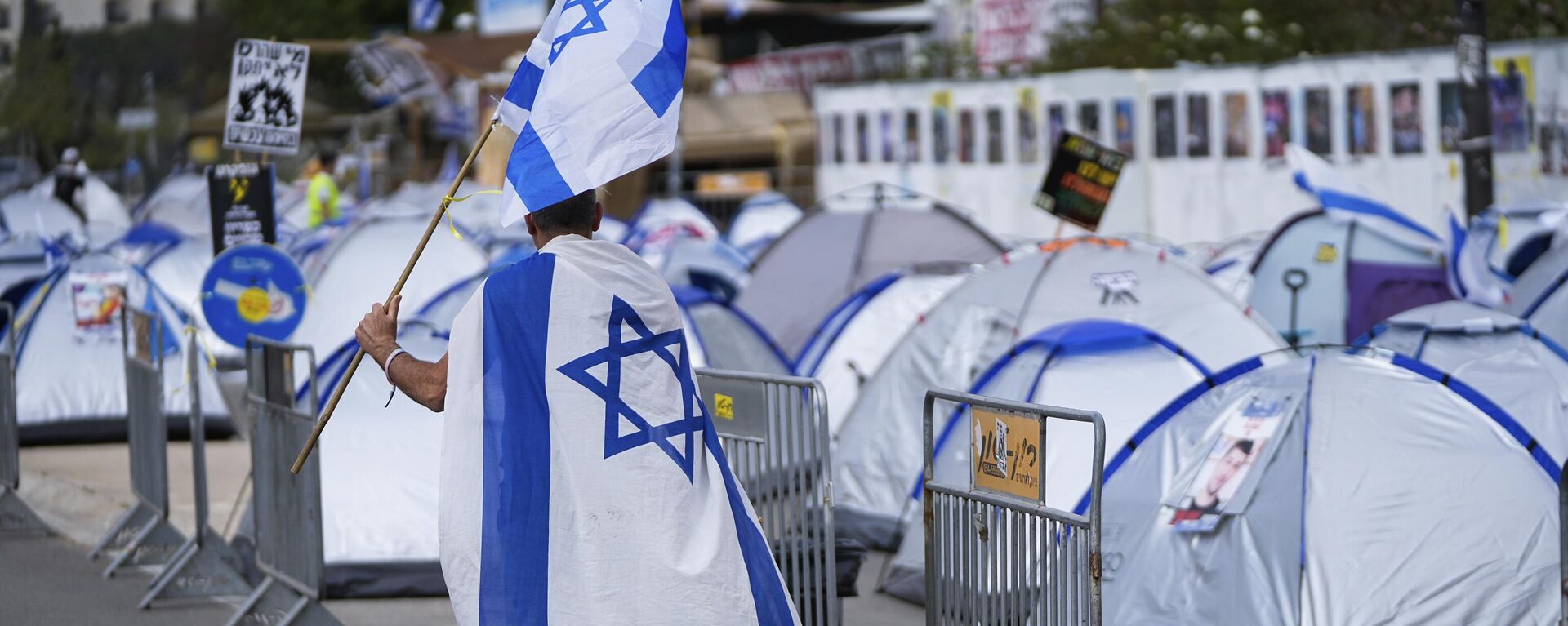 مردی با پرچم اسرائیل از کنار اردوگاهی می‌گذرد که توسط مردم معترض به دولت بنیامین نتانیاهو، نخست‌وزیر اسرائیل و خواهان آزادی گروگان‌هایی که در نوار غزه توسط شبه‌نظامیان حماس در خارج از کنست، پارلمان اسرائیل، در اورشلیم بازداشت شده‌اند، ایجاد شده است - اسپوتنیک بین‌المللی , 1920, 2024/04/19