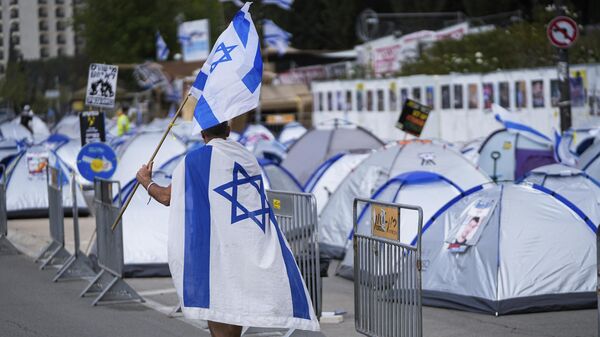 A man walks with the Israeli flag past a camp set up by people who are protesting against Israeli Prime Minister Benjamin Netanyahu's government and call for the release of hostages held in the Gaza Strip by the Hamas militant group near the Knesset, Israel's parliament, in Jerusalem - Sputnik International
