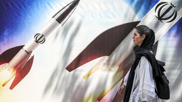 (FILES) A woman walks past a banner depicting launching missiles bearing the emblem of the Islamic Republic of Iran in central Tehran on April 15, 2024. - Sputnik International