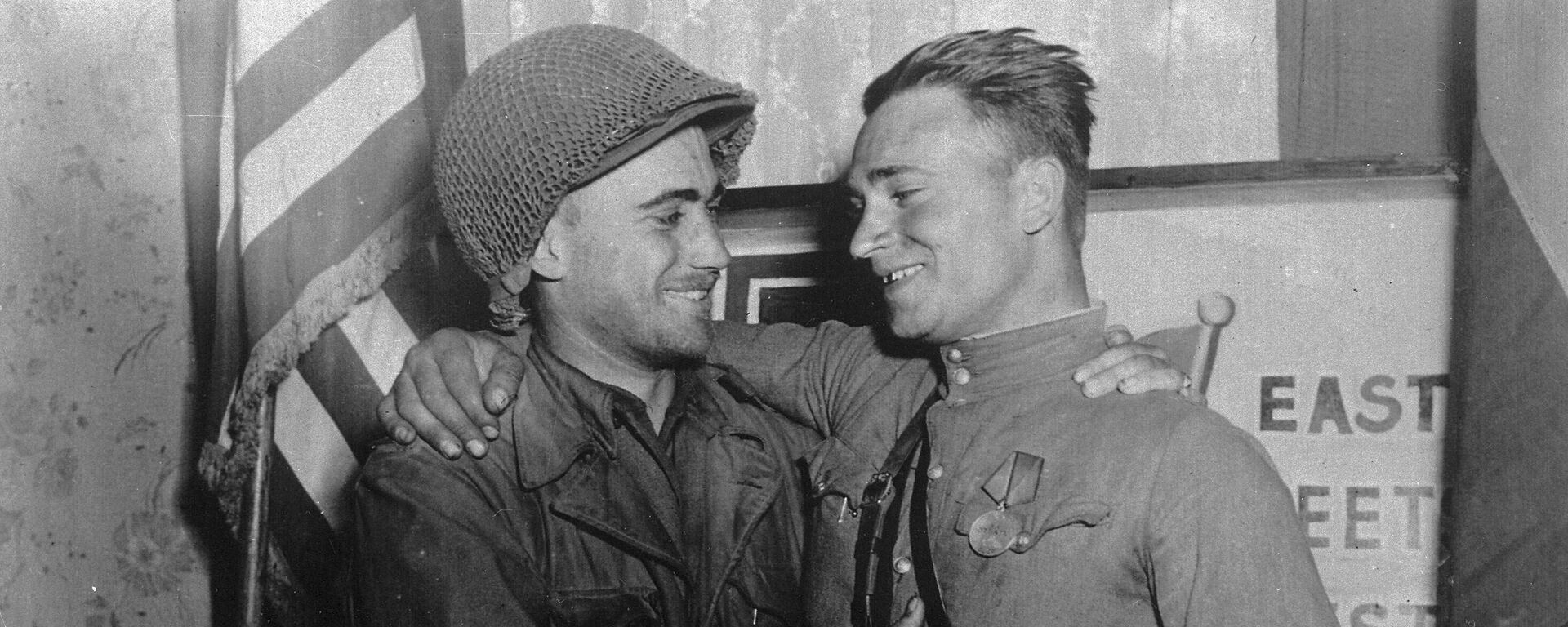 2nd Lt. William Robertson and Lt. Alexander Sylvashko standing in front of a sign that reads East Meets West symbolizing the historic meeting of the Soviet and US Armies - Sputnik International, 1920, 25.04.2024