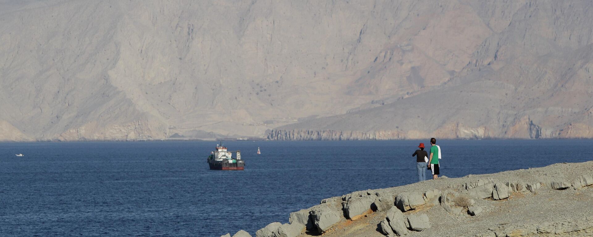 A tourist couple watches the mountains in south of the Strait of Hormuz as the trading dhows and ships are docked on the Persian Gulf waters near the town of Khasab, in Oman - Sputnik International, 1920, 17.04.2026