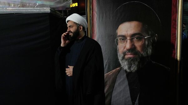 A cleric speaks on his cellphone next to a portrait of Iran's Supreme Leader Ayatollah Mojtaba Khamenei during a pro-government gathering near the residence where his father, former Supreme Leader Ayatollah Ali Khamenei, was killed during US and Israeli strikes on Feb. 28, in Tehran, Iran, Monday, May 18, 2026. - Sputnik International