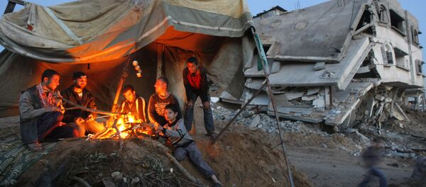 Palestinians sit around a fire under the cover of a tent on the ruins of their home Palestinians sit around a fire under the cover of a tent on the ruins of their home - Sputnik International