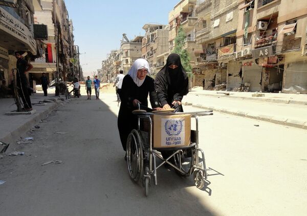 Residents of Syria's Yarmuk Palestinian refugee camp, south of Damascus, use a wheelchair to carry a box of goods distributed by the United Nations Relief and Works Agency (UNRWA) on July 17, 2014 - Sputnik International