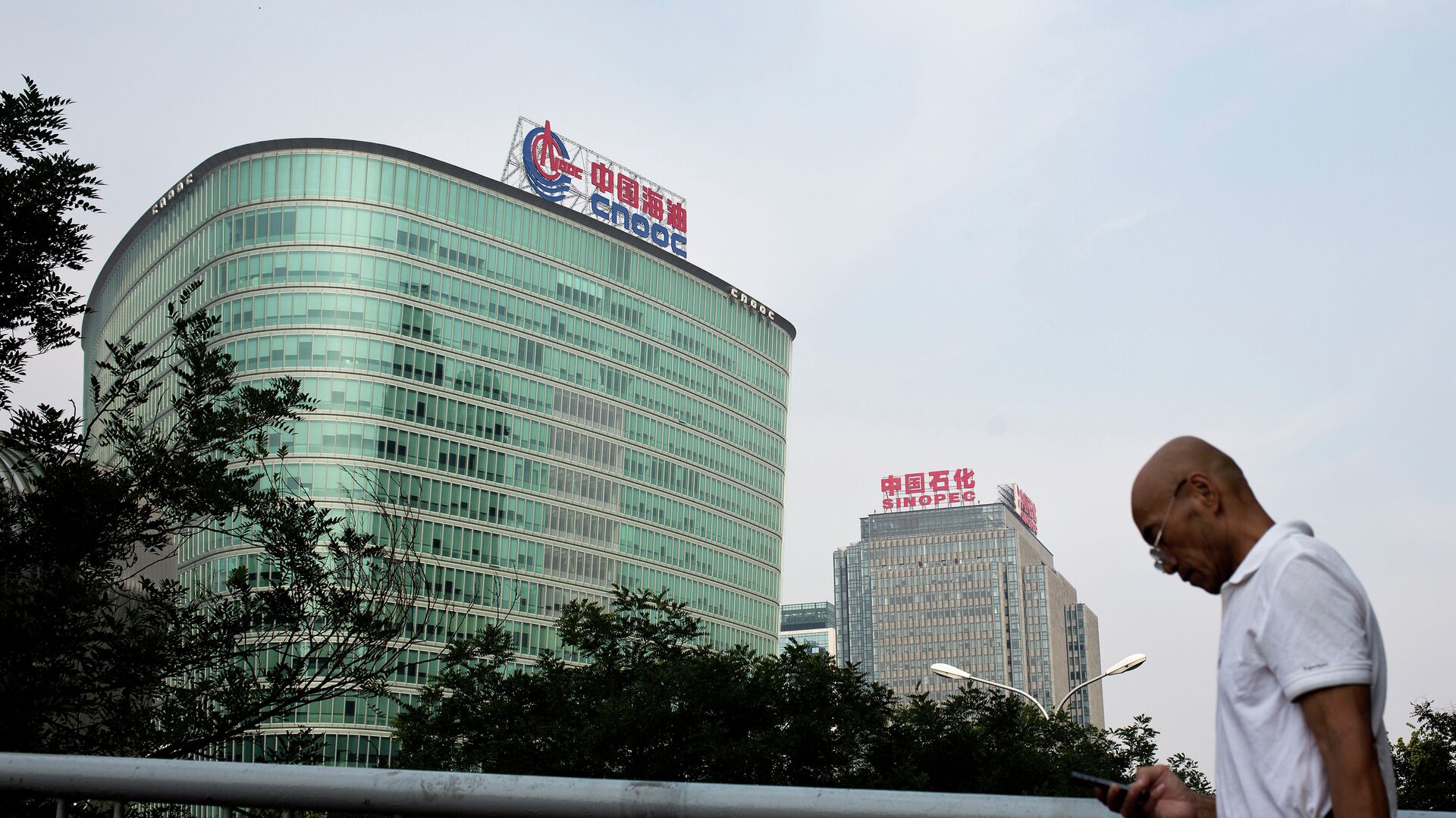 A man checks his mobile phone as he walks by buildings of China's state-owned companies, China National Offshore Oil Corp. (CNOOC), left, and China Petroleum & Chemical Corp. (Sinopec), in Beijing Monday, Sept. 14, 2015 - Sputnik International, 1920, 26.03.2023