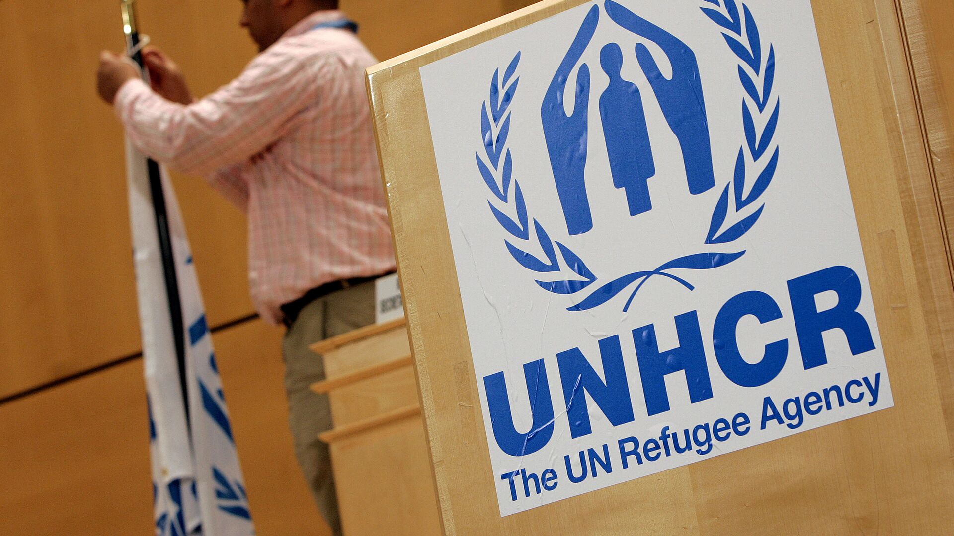 A staff installs a UNHCR flag close to a logo of UN High Commissioner for Refugees (UNHCR) prior to the opening of a two-day United Nations conference trying to boost support for Iraqis who have fled violence, 17 April 2007 at the UN Office in Geneva - Sputnik International, 1920, 19.02.2022