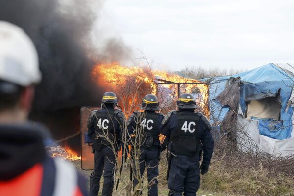 French CRS riot police watch as smoke and flames billow from a burning makeshift shelter. French CRS riot police watch as smoke and flames billow from a burning makeshift shelter. - Sputnik International
