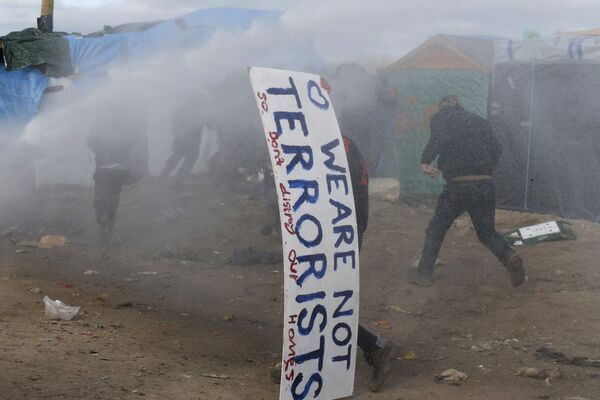 A man takes cover with a sign that reads, We Are Not Terrorists - So Don't Destroy Our Homes as water canon and tear gas push back migrants. A man takes cover with a sign that reads, We Are Not Terrorists - So Don't Destroy Our Homes as water canon and tear gas push back migrants. - Sputnik International