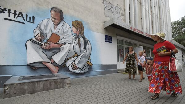 Women with children walk past graffiti showing Russian President Vladimir Putin, dressed in a Judogi and sitting next to a girl, on the front of a children's clinic in Yalta, Crimea, Wednesday, Aug. 19, 2015 - Sputnik International
