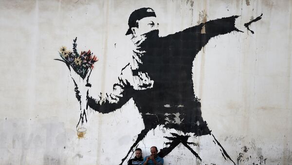 Two men are sitting in front of a famous graffiti of British street artist Banksy, painted on a wall of a gas station in the West Bank city of Bethlehem on 16 December 2015.  - Sputnik International