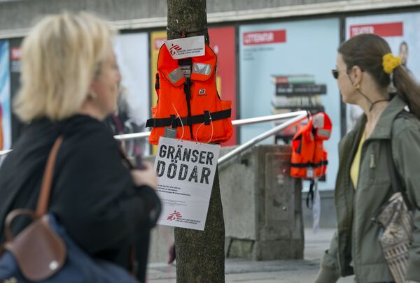 Women pass by a life vest and a placard reading Borders kill hanging on a tree in Stockholm as part of an action by Swedish branch of humanitarian NGO Doctors without borders, in solidarity with migrants seeking asylum in Europe after fleeing their home countries on September 14, 2015 - Sputnik International