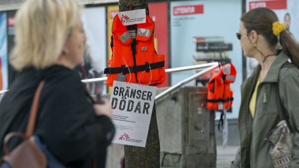 Women pass by a life vest and a placard reading Borders kill hanging on a tree in Stockholm as part of an action by Swedish branch of humanitarian NGO Doctors without borders, in solidarity with migrants seeking asylum in Europe after fleeing their home countries on September 14, 2015 - Sputnik International