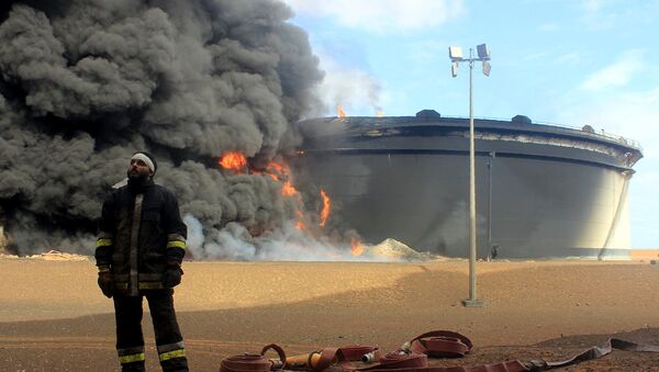 A Libyan fireman stands in front of smoke and flames rising from an oil storage tank at an oil facility in northern Libya's Ras Lanouf region on January 23, 2016, after it was set ablaze earlier in the week following attacks launched by Islamic State (IS) group jihadists to seize key port terminals. - Sputnik International