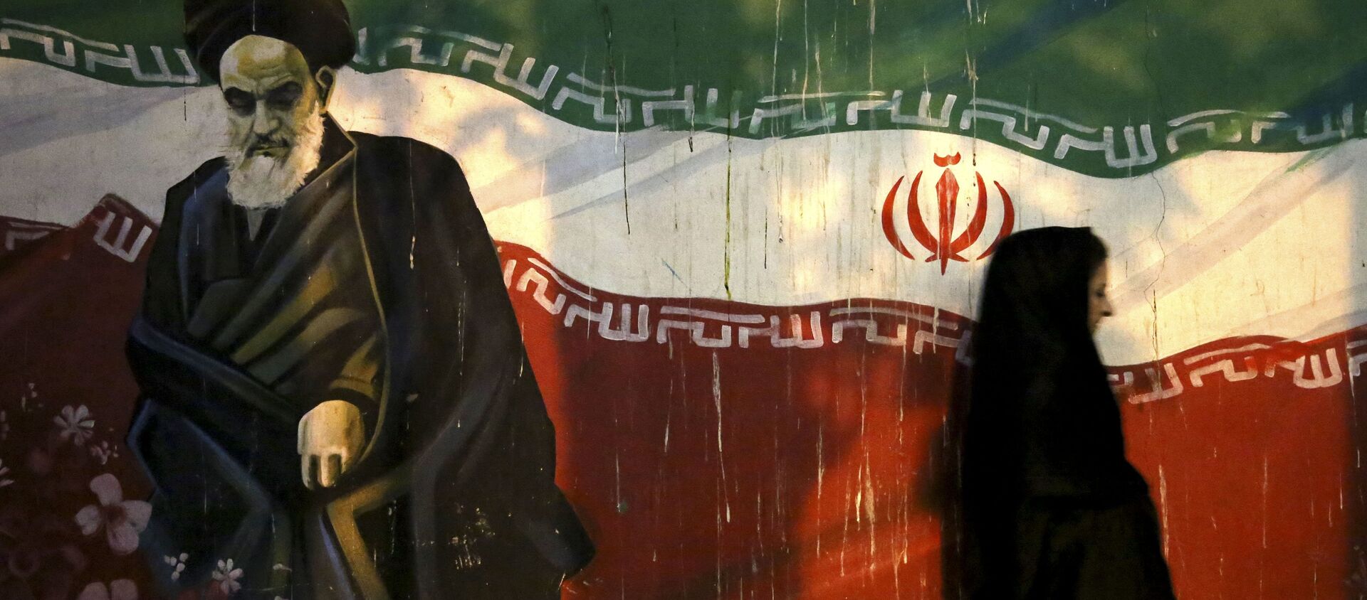 veiled Iranian woman walks past a mural depicting the late revolutionary founder Ayatollah Khomeini, and national Iranian flag, painted on the wall of the former U.S. Embassy, in Tehran, Iran - Sputnik International, 1920, 21.02.2021