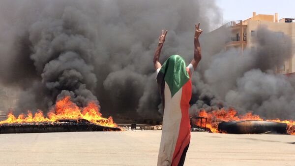 A protester wearing a Sudanese flag flashes the victory sign in front of burning tires and debris on road 60, near Khartoum's army headquarters. - Sputnik International