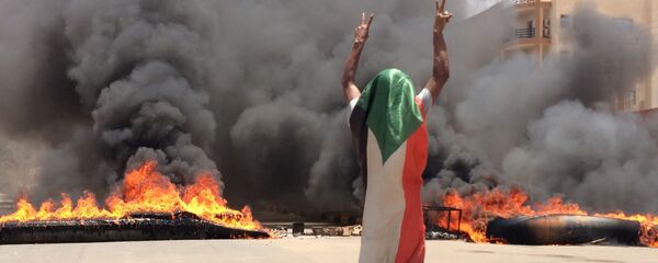 A protester wearing a Sudanese flag flashes the victory sign in front of burning tires and debris on road 60, near Khartoum's army headquarters. A protester wearing a Sudanese flag flashes the victory sign in front of burning tires and debris on road 60, near Khartoum's army headquarters. - Sputnik International