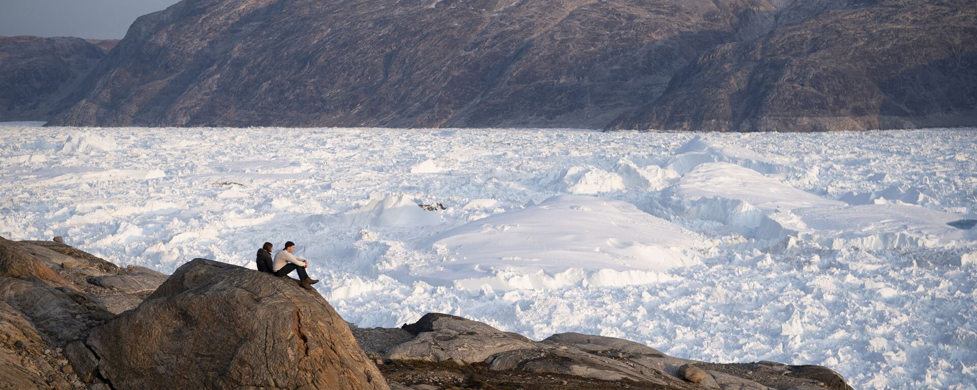 In this Aug. 16, 2019, photo, New York University student researchers sit on a rock overlooking the Helheim glacier in Greenland. - Sputnik International, 1920, 23.01.2025