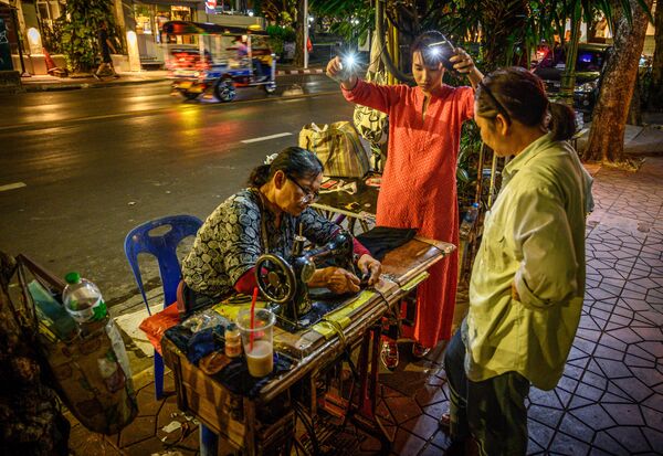 A woman uses the lights of two mobile phones to help a street tailor finish her work on a street in Bangkok on December 3, 2019.  - Sputnik International