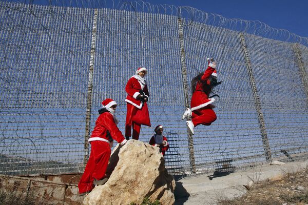 Palestinians wearing Christmas costumes jump off a rock by a barbed-wired section of Israel's separation barrier near the village of Wallajeh, west of the biblical city of Bethlehem in the West Bank - Sputnik International