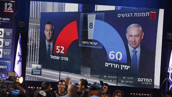 This picture taken on March 2, 2020 shows people standing before a giant screen displaying a broadcast from Israel's Channel 13 with an exit poll prediction after polls closed in the Israeli elections, predicting that Prime Minister Benjamin Netanyahu's Likud party expects to command a 60 seat bloc against 52 for Benny Gantz' Blue and White (Kahol Lavan) electoral alliance, at the Blue and White's headquarters in the coastal city of Tel Aviv. - Sputnik International