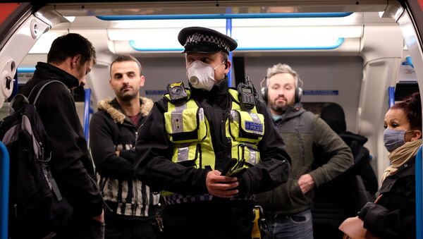 A police officer wearing PPE (personal protective equipment), including a face mask as a precautionary measure against COVID-19, stands with commuters as they travel in the morning rush hour on TfL (Transport for London) London underground Victoria Line trains from Finsbury Park towards central London on May 13, 2020 - Sputnik International