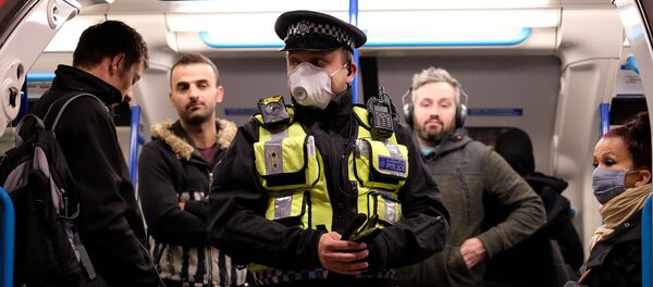 A police officer wearing PPE (personal protective equipment), including a face mask as a precautionary measure against COVID-19, stands with commuters as they travel in the morning rush hour on TfL (Transport for London) London underground Victoria Line trains from Finsbury Park towards central London on May 13, 2020 - Sputnik International