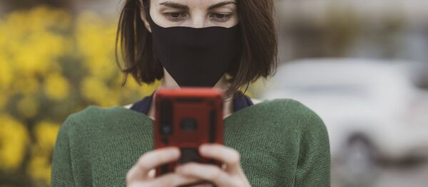 A woman wearing a face mask holds up her mobile phone - Sputnik International