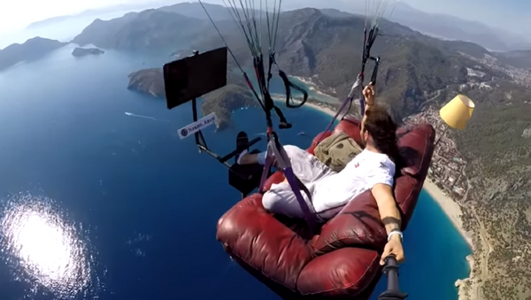 A screenshot of a video of Hasan Kaval, a Turkish paragliding instructor, watching TV while flying over water on a couch attached to a paraglider. - Sputnik International